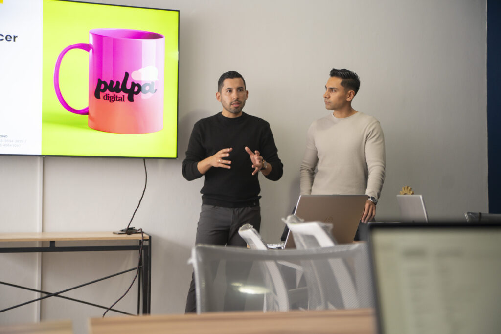 Es la fotografía de dos de los expertos de marketing digital de la agencia Pulpa Digital, dando una conferencia al equipo de la promotoría de Abundance. Al lado de ellos está una pantalla donde se puede ver una taza de color rosa con el logo de Pulpa Digital, sobre un fondo verde.